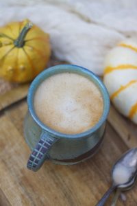 Blue mug sitting on a wooden cutting boards surrounded by small pumpkins, white cloth, and a metal spoon.